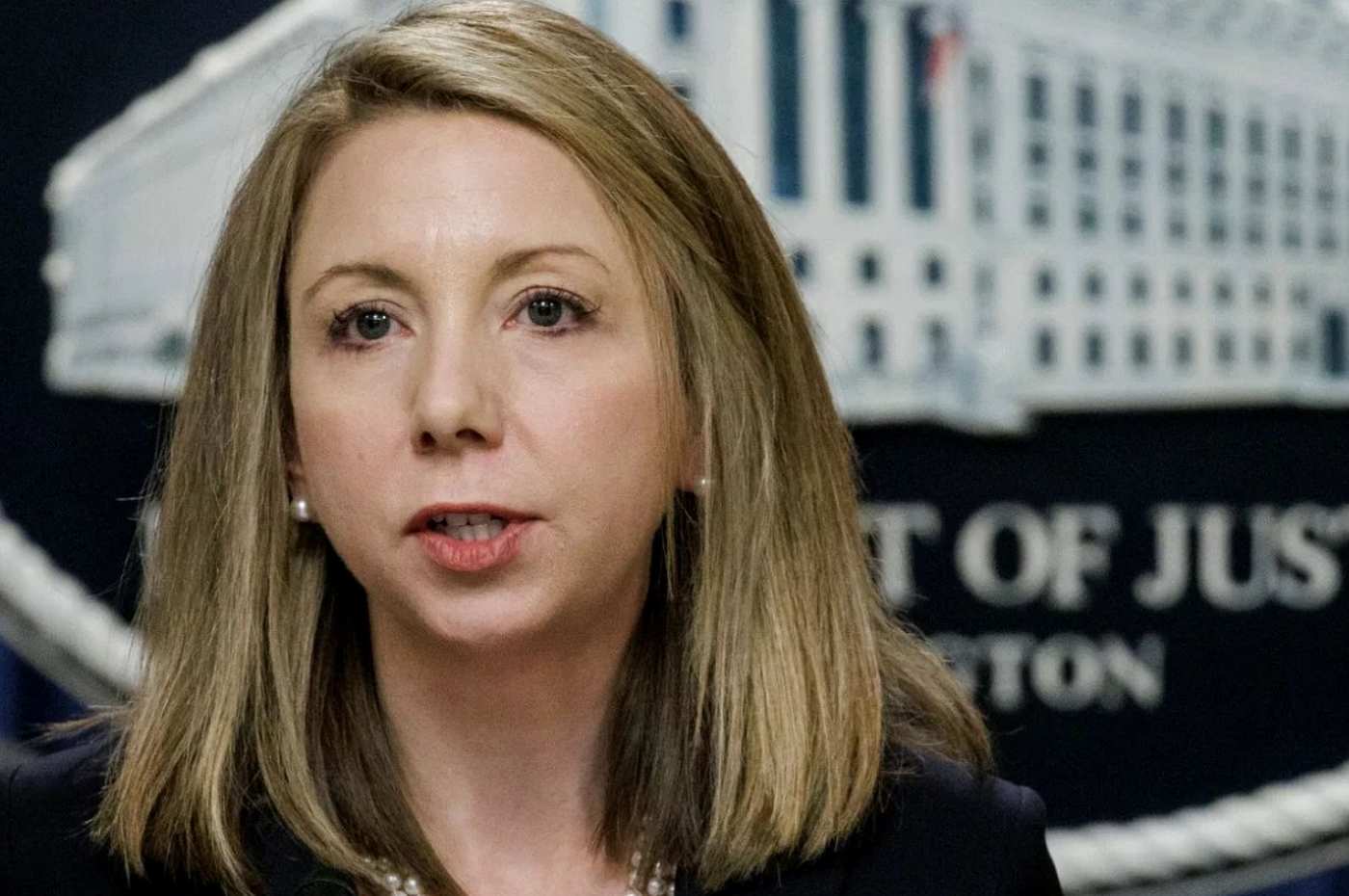 department of Justice official speaking at podium with DOJ seal visible in background