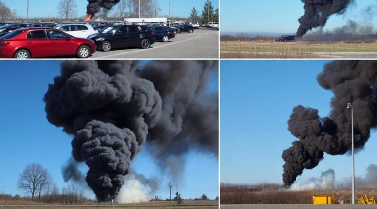 thick black smoke plume rising from industrial fire, visible from parking lot and rural
