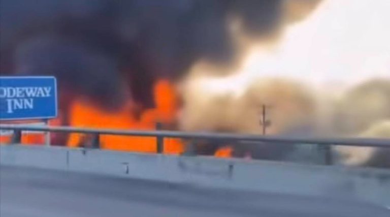 intense orange flames and thick black smoke rising behind highway guardrail near blue