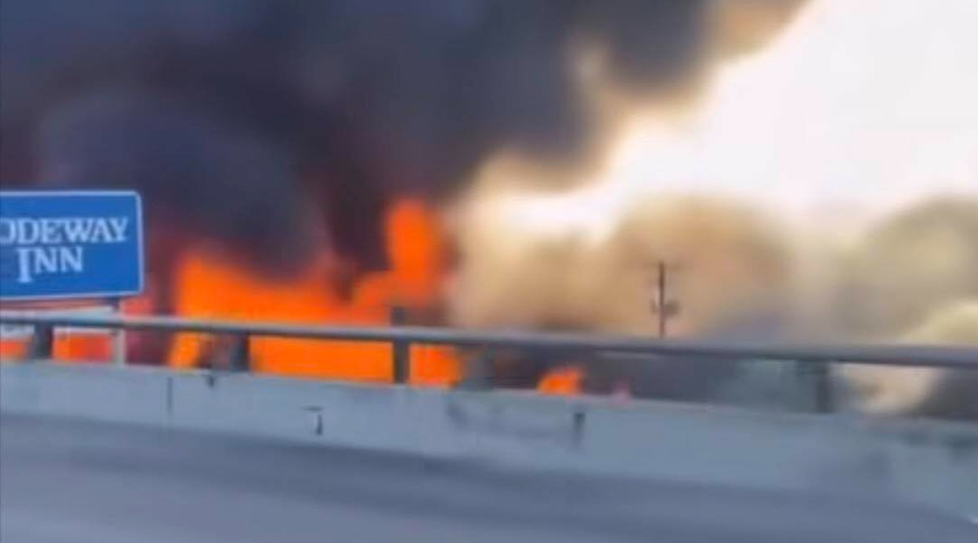 intense orange flames and thick black smoke rising behind highway guardrail near blue