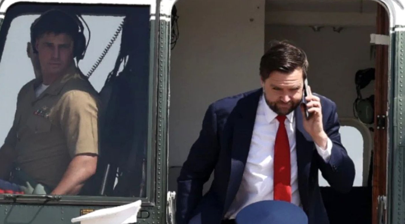 JD Vance in navy suit and red tie exits helicopter while talking on phone, pilot visible